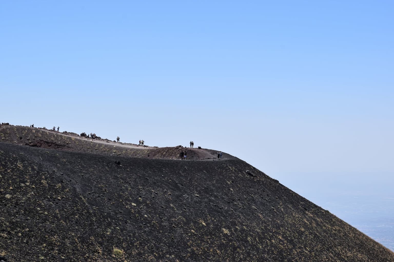 A small group of hikers walking a ridge of black volcanic ash on Mount Etna's upper slope, the scale of the bare mountainside behind them.