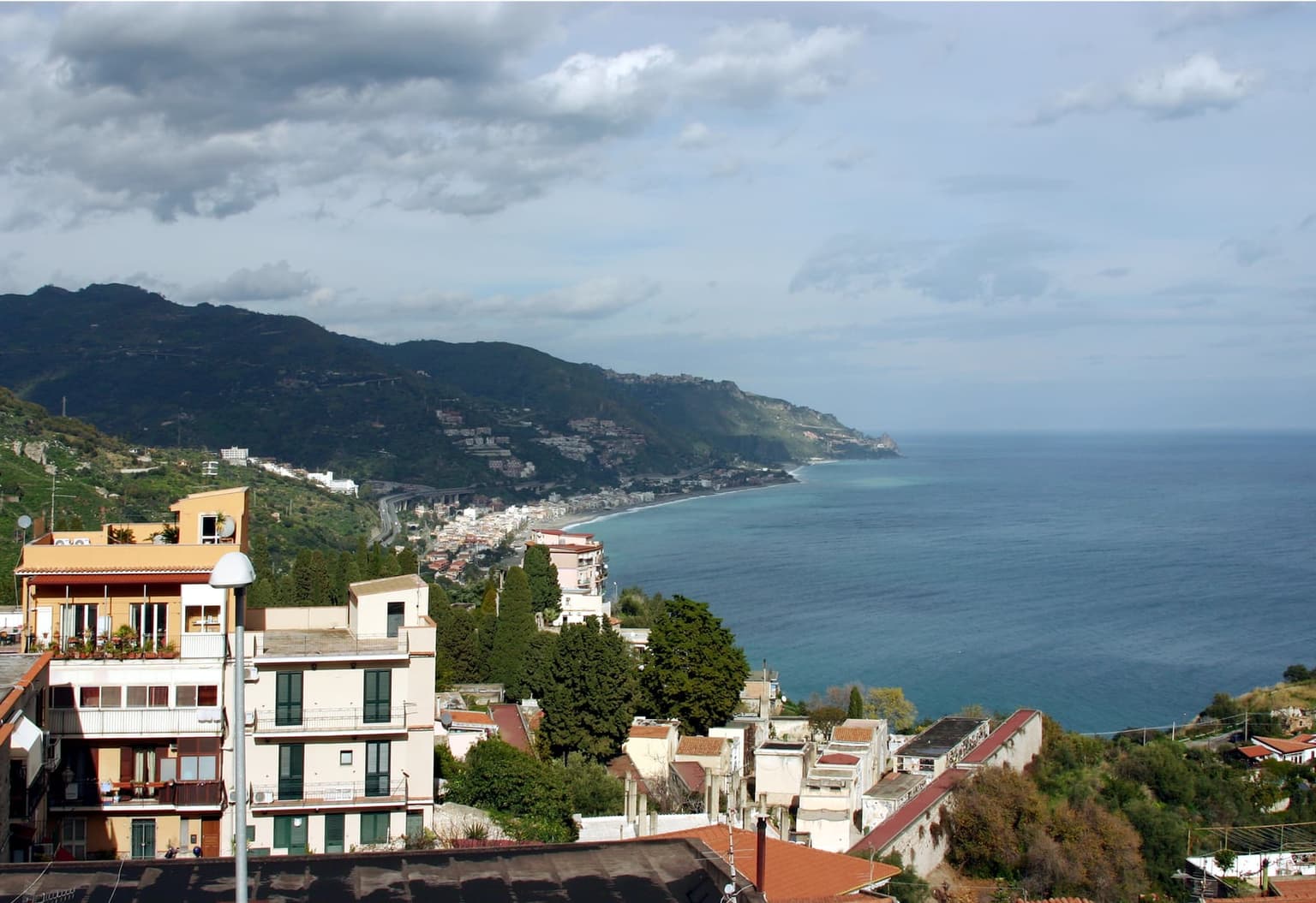 An elevated coastal view south from a hillside town, showing apartment buildings in the foreground and a lower town along a curving bay in the distance.