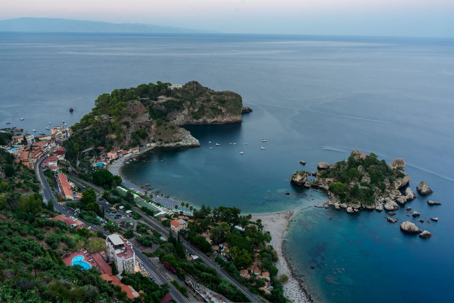 Aerial view at dusk of the Isola Bella cove, the narrow pebble isthmus connecting the wooded islet to the mainland beach, boats moored in the bay.