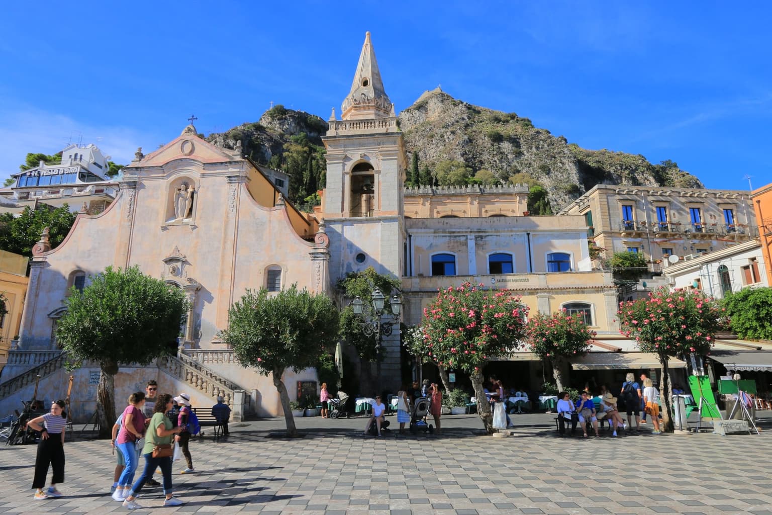 Piazza IX Aprile's checkerboard-paved terrace with visitors gathered near a Baroque church and bell tower, a rocky hillside rising behind.