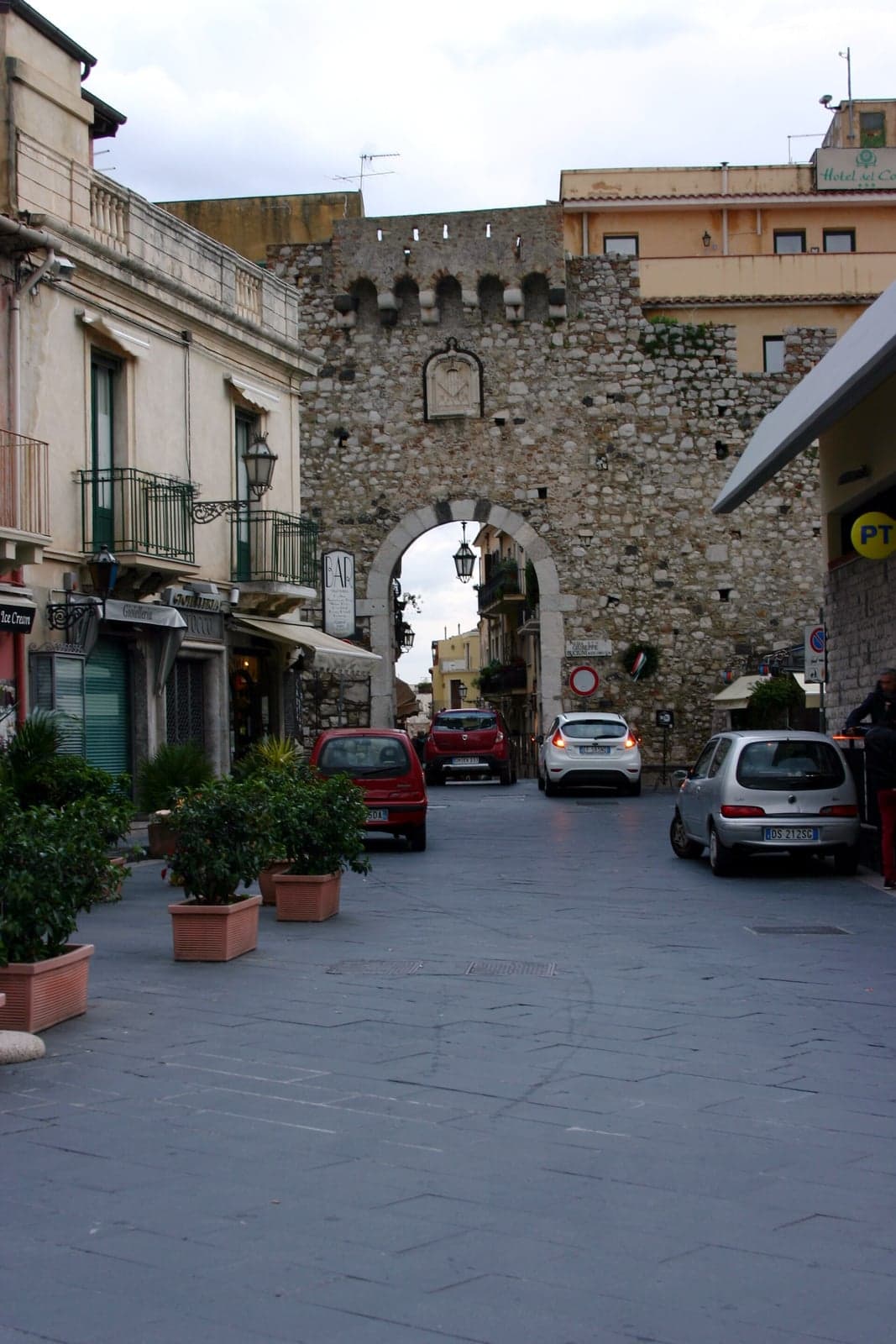 A stone city-wall arch opening onto a paved street lined with shops, several cars parked along the curb beside it.