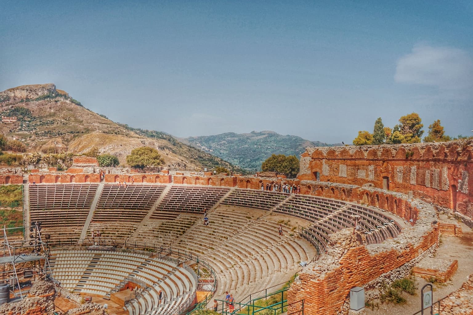 The Teatro Antico's curved stone seating bowl seen from the upper rows, mostly empty, a scatter of visitors on the tiered steps.