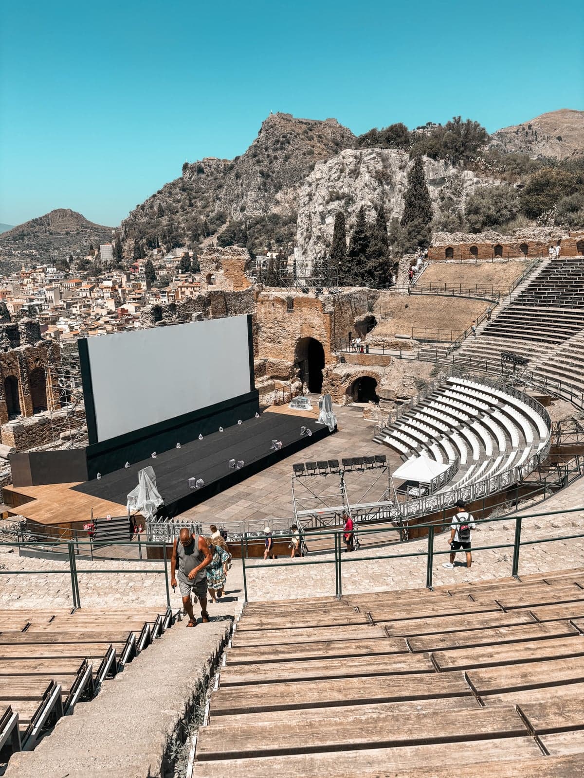 A modern concert stage with a large screen and lighting rig set up inside the ancient theater's cavea, temporary white seating rows fitted among the ruins.
