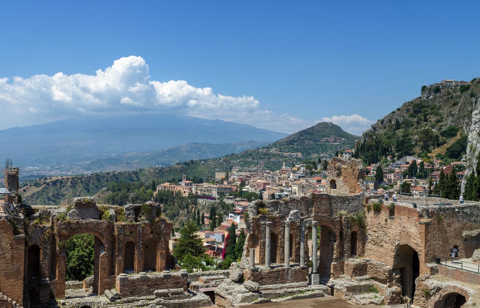 The Teatro Antico's ruined brick stage wall and standing columns in the foreground, framing a view of Taormina's rooftops and snow-capped Mount Etna beyond.
