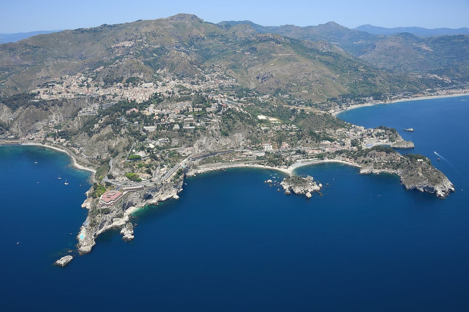 Aerial view of Taormina spread across its hillside terrace above the Ionian Sea, with the wooded islet of Isola Bella and the coastline curving away on both sides.