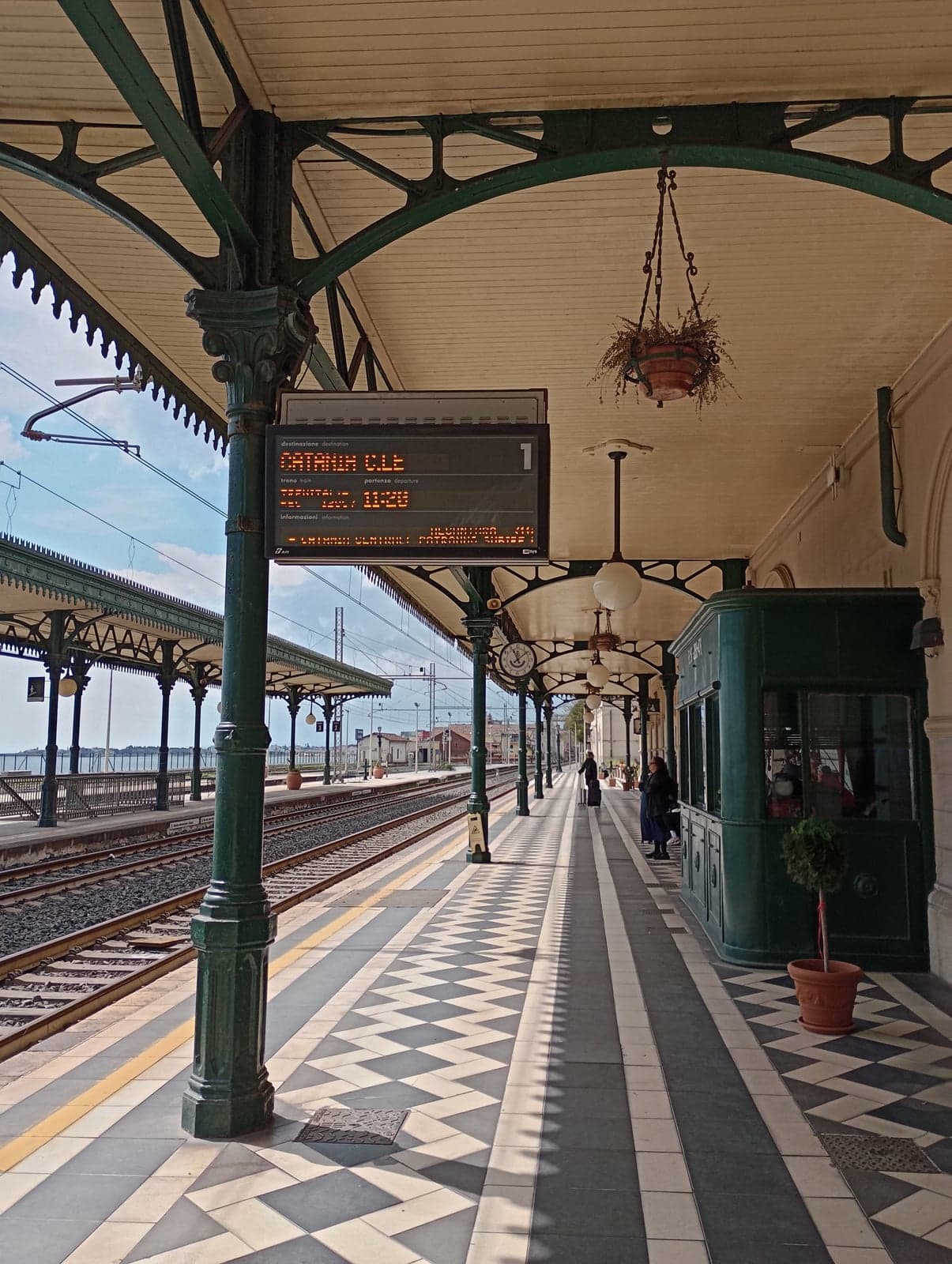 Ornate green ironwork canopy over platform 1 at Taormina-Giardini station, a checkerboard-tiled floor, a hanging flower basket, and an illuminated departure board reading "CATANIA C.LE" with an 11:29 departure, the sea visible past the platforms in the distance.