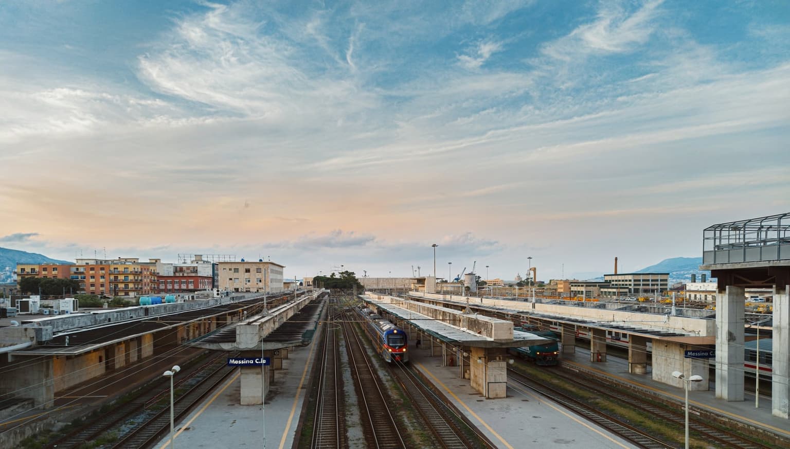 Elevated view over Messina Centrale station at dusk, multiple platforms and tracks under a pastel sky, a blue "Messina C.le" sign on the platform, a train arriving on the near track.