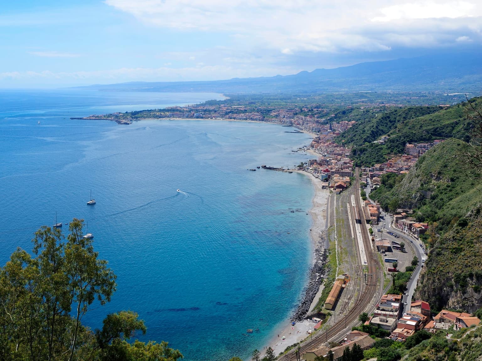 Aerial view down a hillside onto the curving bay of Giardini Naxos, Taormina-Giardini railway station's platforms and tracks running along the shoreline between a green cliff and turquoise water, sailboats anchored offshore, the town and distant mountains wrapping the coastline beyond.