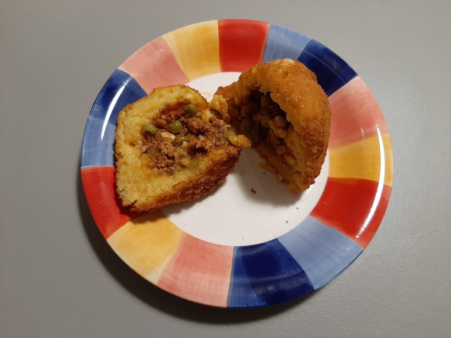 A round Sicilian arancina cut in half on a plate, showing the meat ragù and peas filling inside its fried rice shell