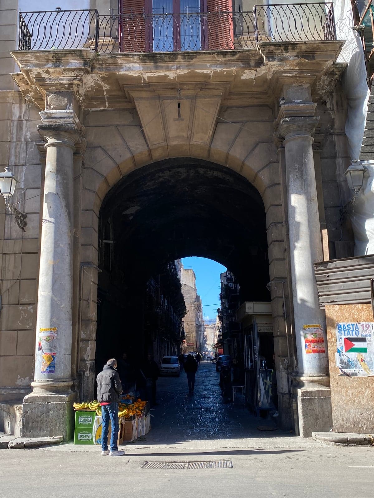 A stone archway opening onto a Palermo market street, produce crates stacked at its base and market stalls visible in the street beyond