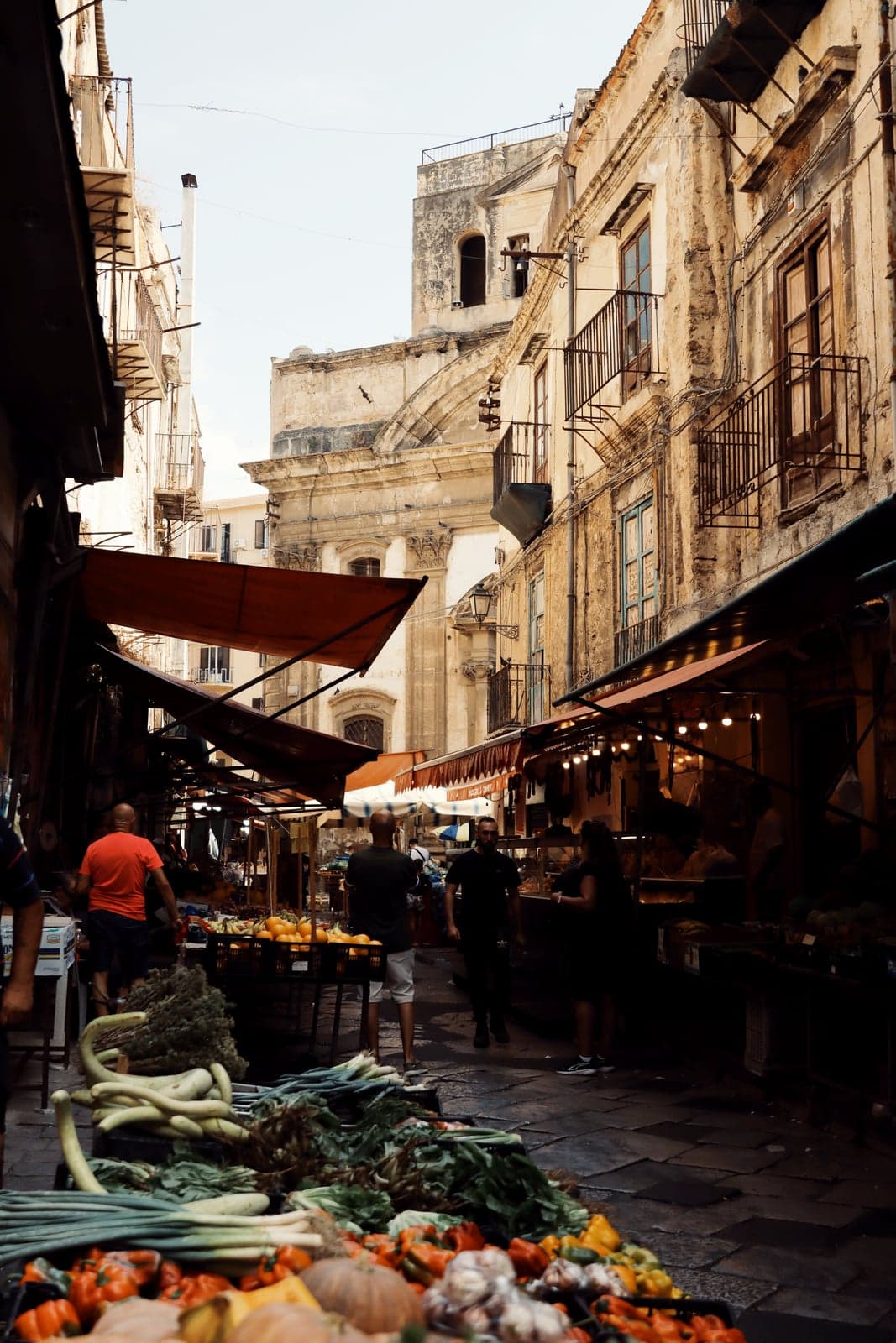 A narrow Ballarò market alley with striped awnings, fresh vegetables piled in the foreground, and shoppers moving between the stalls