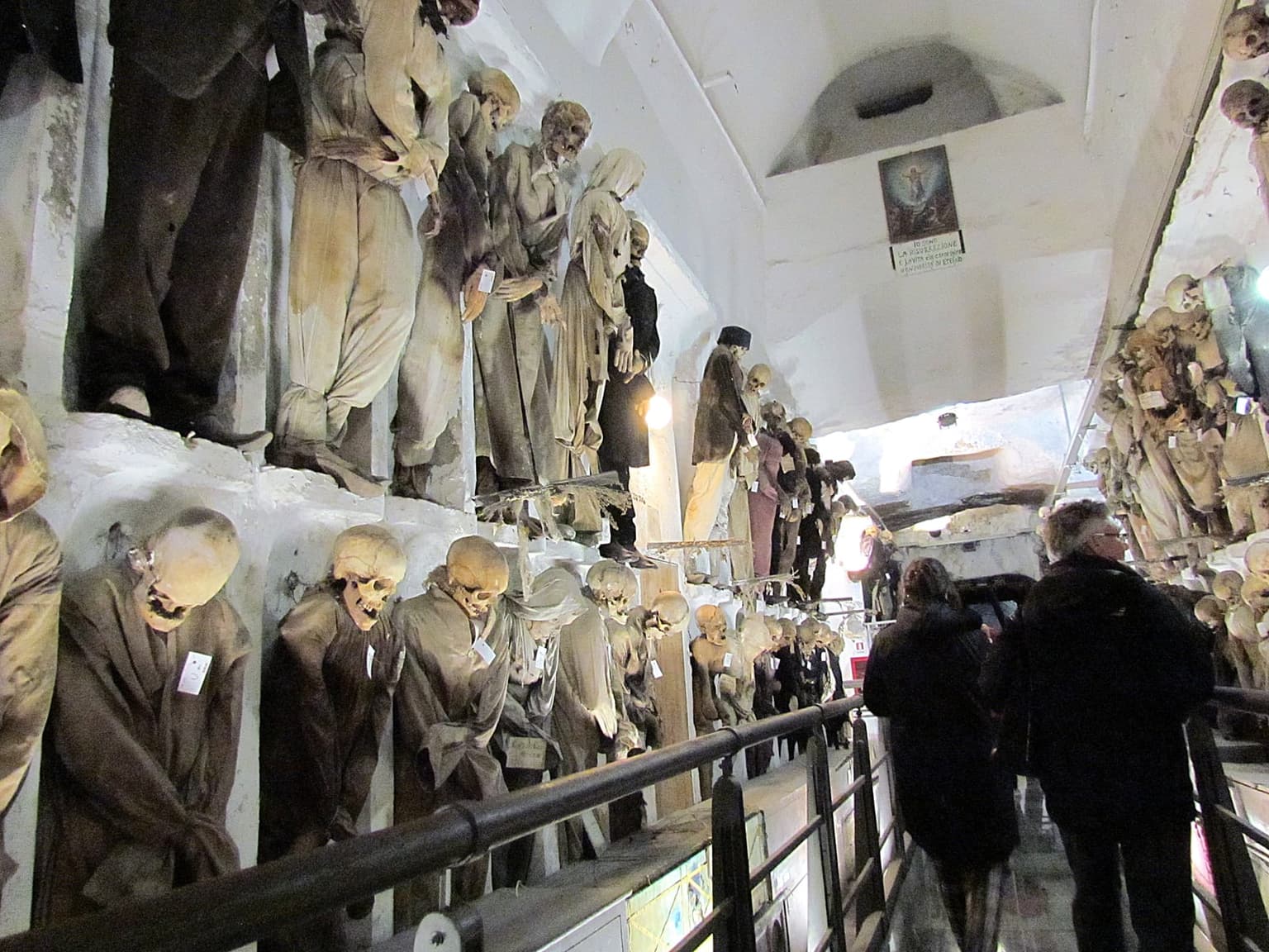 A corridor of the Capuchin Catacombs lined with standing mummified figures in period dress, dust visible on the fabric, receding into the passage