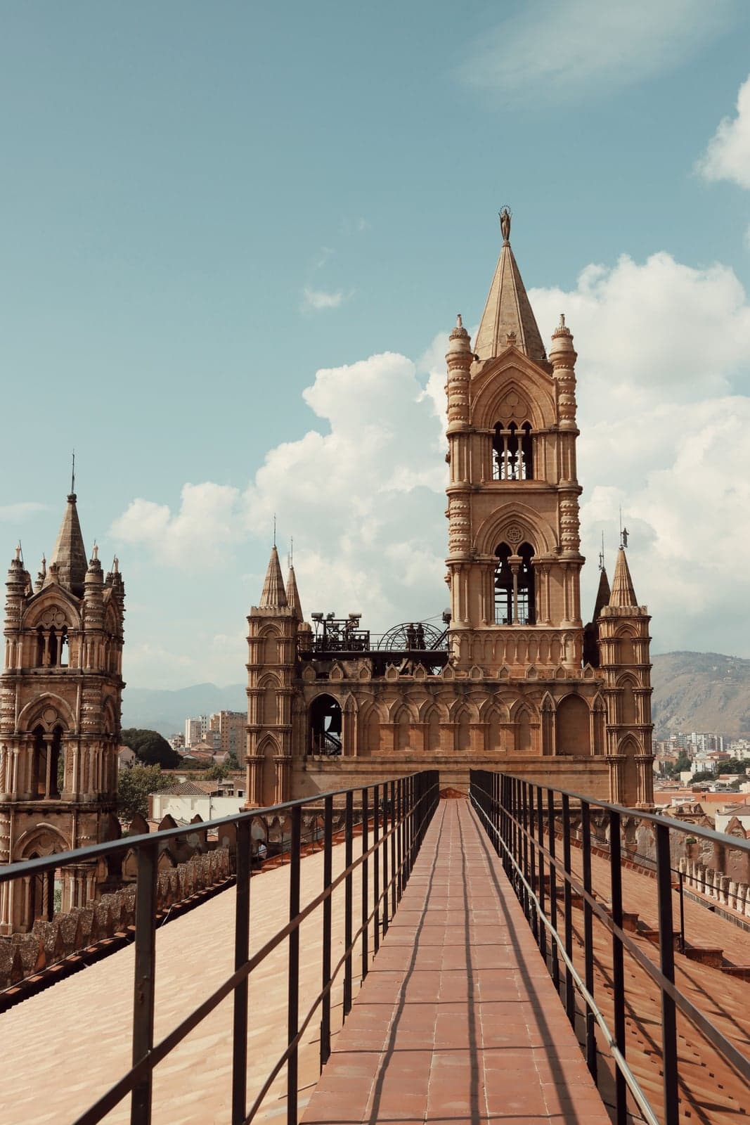 A tiled rooftop walkway between Palermo Cathedral's Gothic spires, railing on both sides, with the city's rooftops and the surrounding mountains beyond