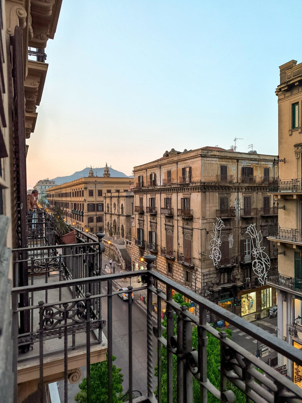 A wide Palermo street at dusk seen from a balcony above, buildings lit and a car passing below, an ordinary evening scene