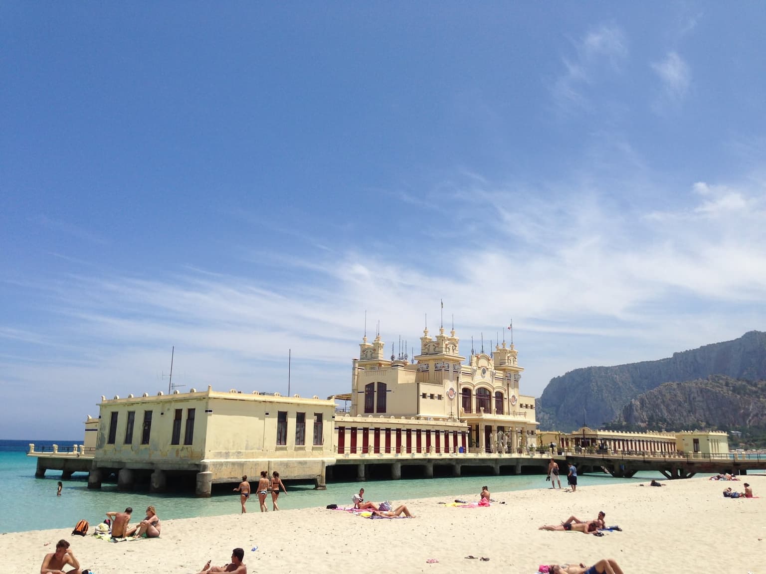 Mondello's Liberty-style bathing pavilion standing on pilings over turquoise water, a rocky headland behind it and people on the sand in front