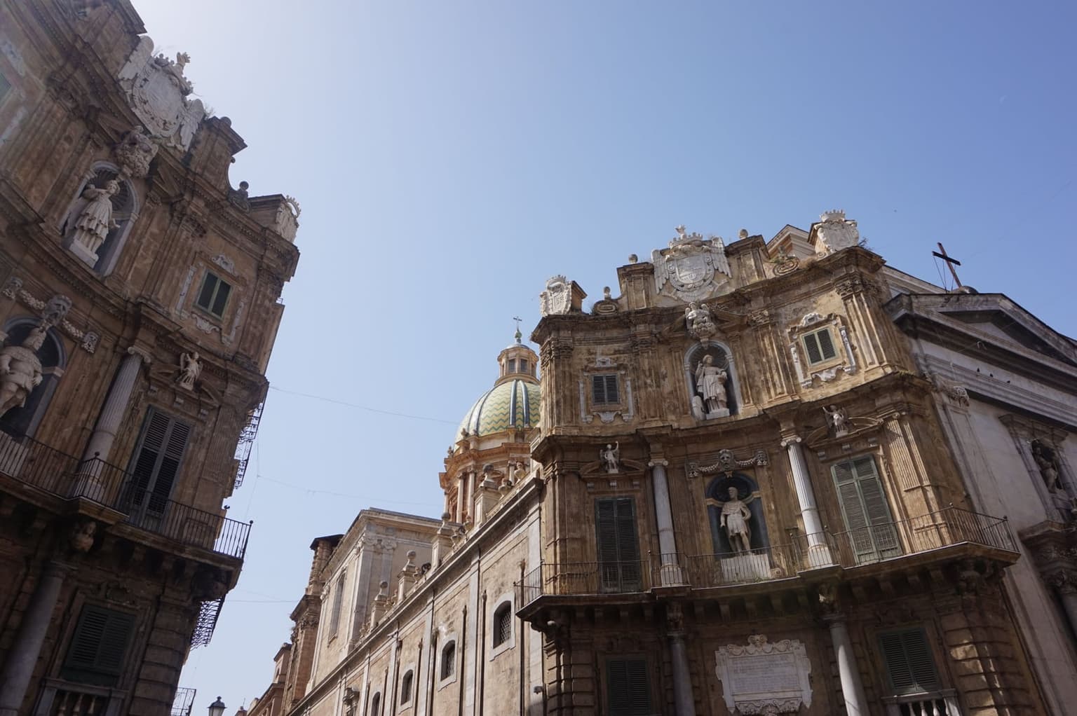 Looking up at the curved Baroque facades of Palermo's Quattro Canti, a green-and-gold church dome visible between the buildings under a clear sky