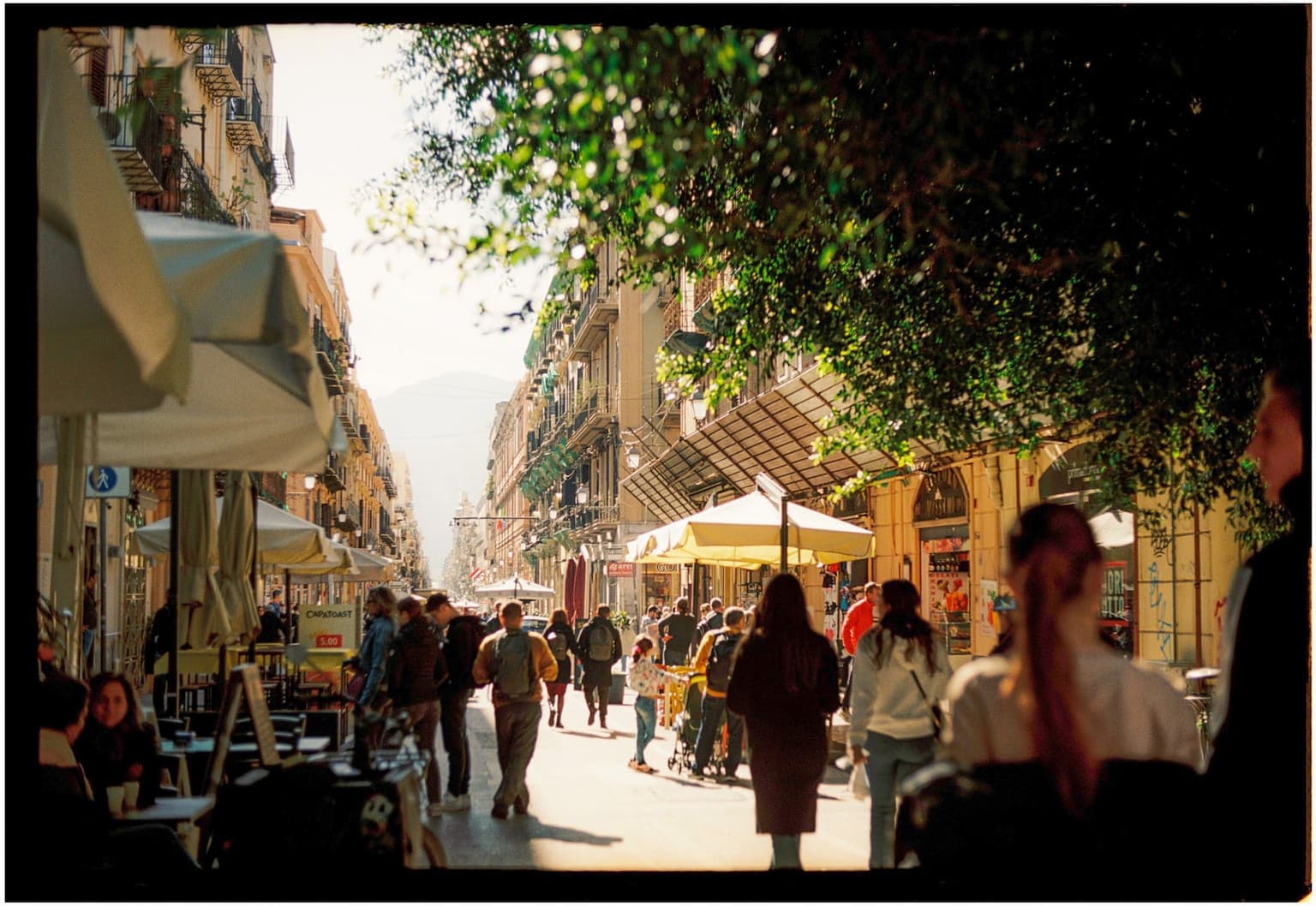 Via Maqueda's pedestrian stretch in Palermo, filled with people walking under café umbrellas and iron balconies on aging ochre facades, no traffic in the roadway