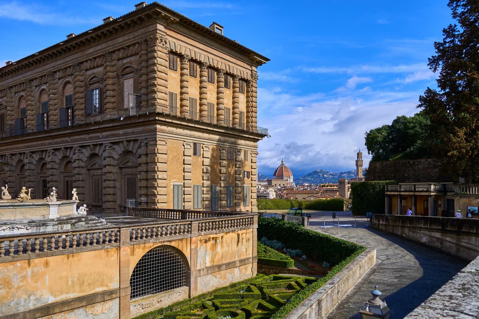 The stone corner of the Pitti Palace seen from the Boboli Gardens, above a sunken garden of clipped hedges, with the cathedral dome and the Palazzo Vecchio tower beyond