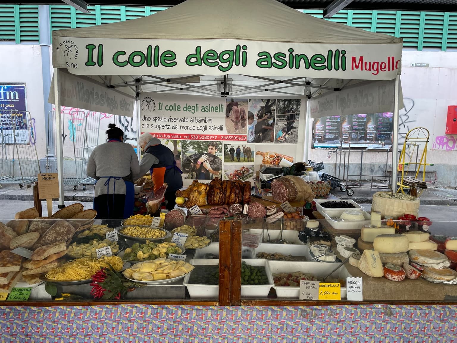 A Mugello farm stall at a covered market, its counter full of bread, fresh pasta, olives, porchetta, salami and cheeses with handwritten price tags