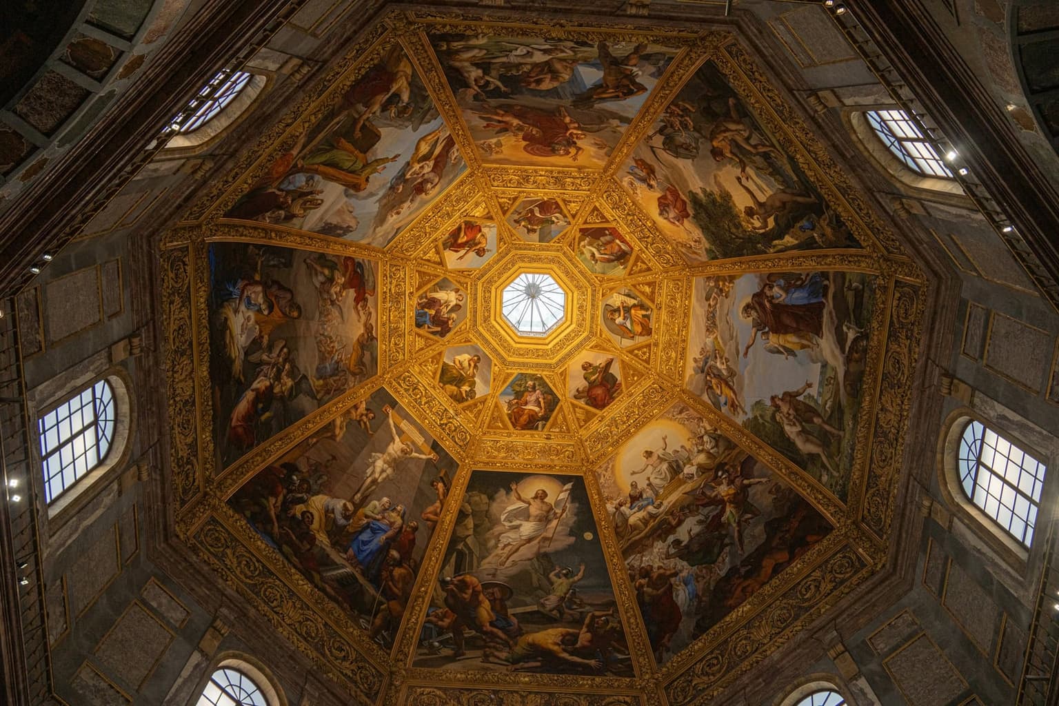 Looking straight up into an octagonal chapel dome, eight painted Bible scenes in gilded frames ringing a small glass lantern