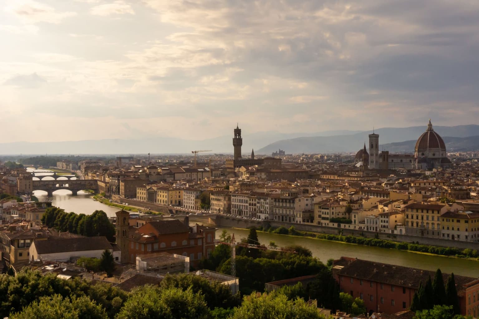 Florence from the hill south of the Arno: the river curving past Ponte Vecchio, the Palazzo Vecchio tower at center and the cathedral dome and bell tower on the right under heavy clouds