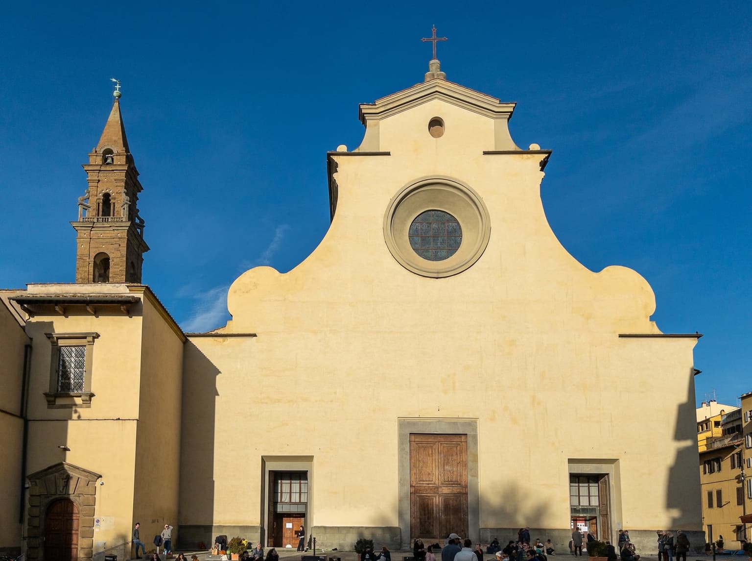 The plain cream-colored front of Santo Spirito church and its brick bell tower, with people sitting on the steps in late-afternoon sun