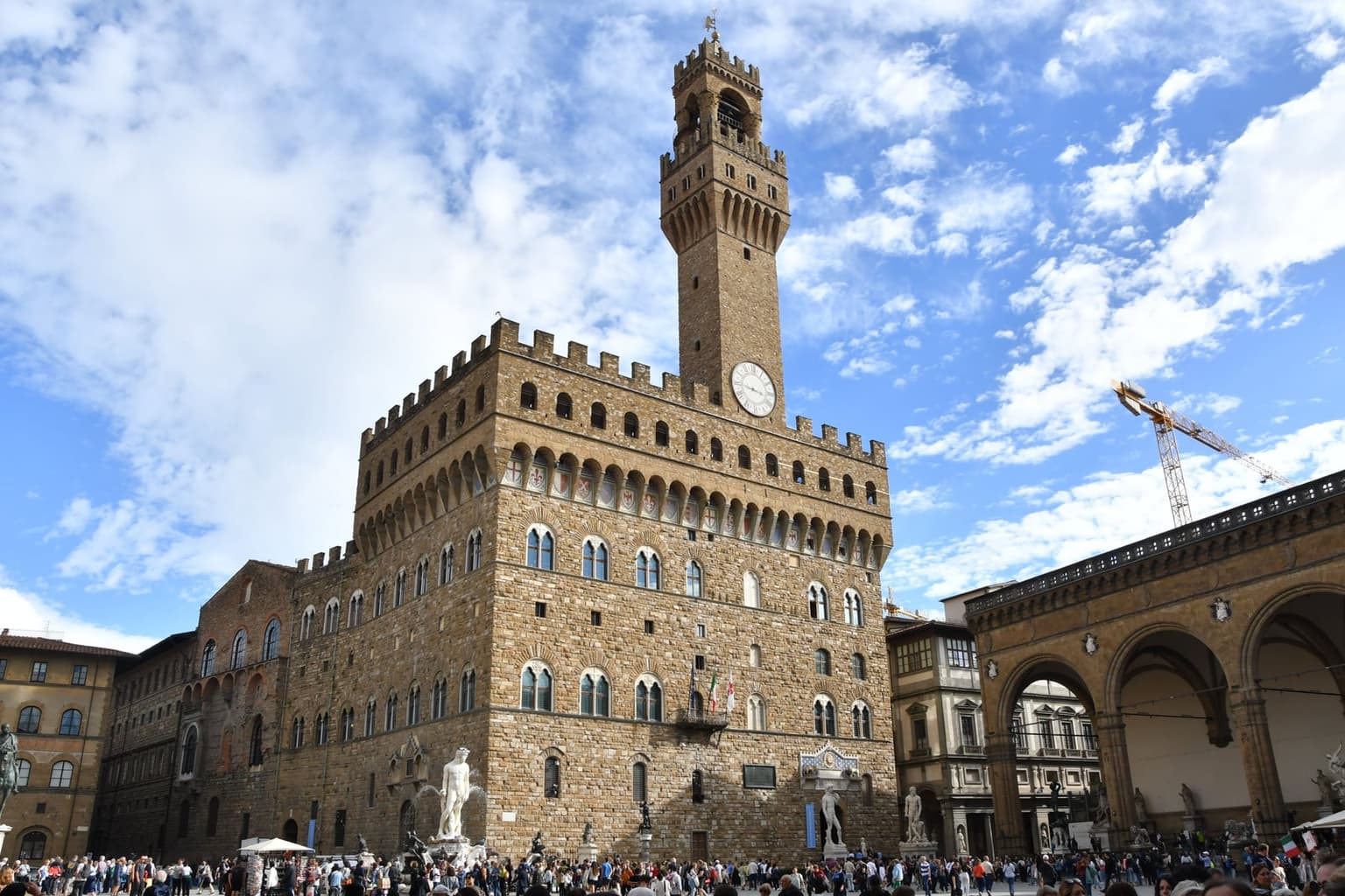 Palazzo Vecchio and its crenellated clock tower above Piazza della Signoria, a marble fountain and statues at its base and a crowd in the square