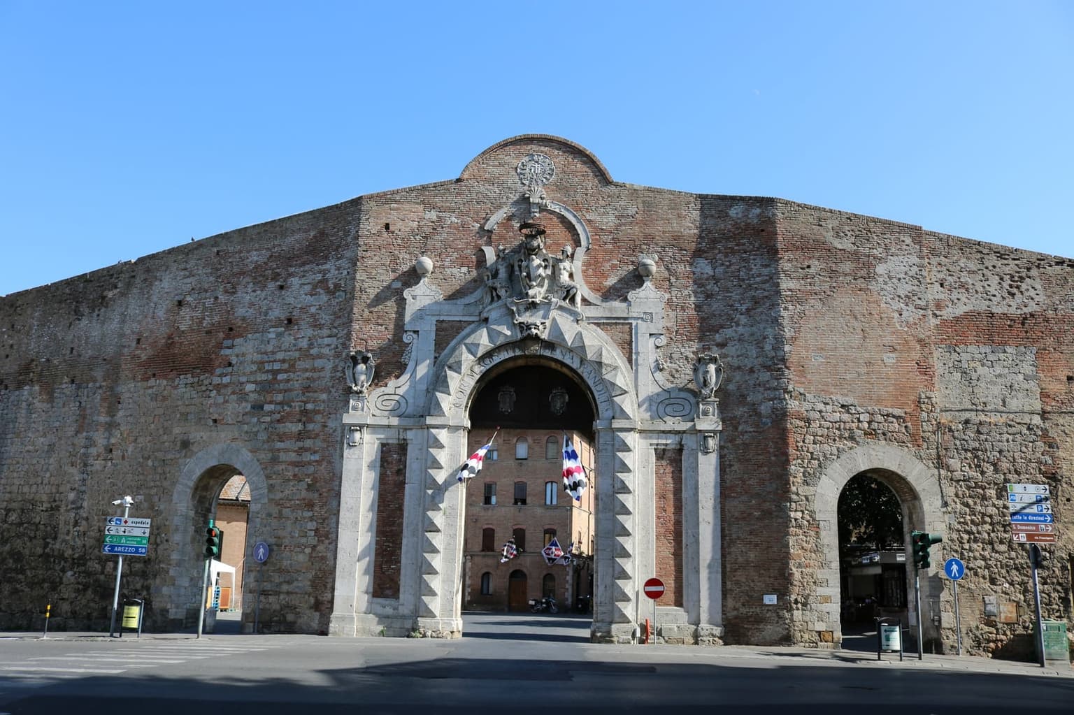 Porta Camollia, a gate in Siena's brick and stone city walls, with a large central arch framed in carved white stone, a smaller arch on each side, and the Latin inscription Cor magis tibi Sena pandit around the main arch.