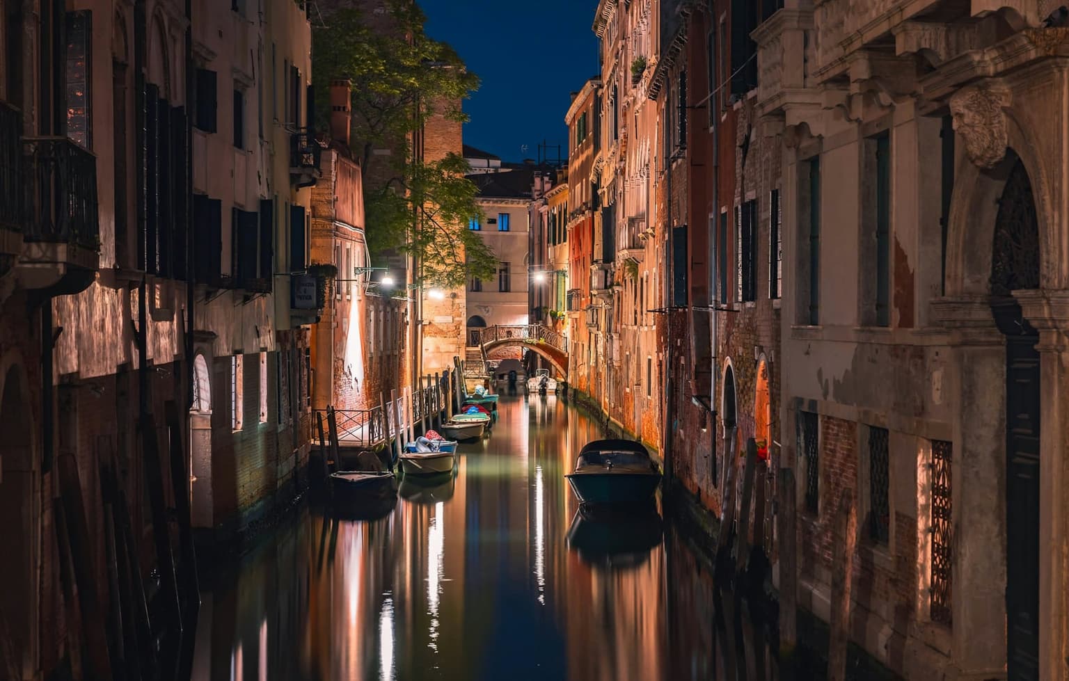 A narrow Venetian canal at night, lamps lighting the old facades and their reflections, small boats moored along the walls and an iron footbridge at the far end, no one in sight