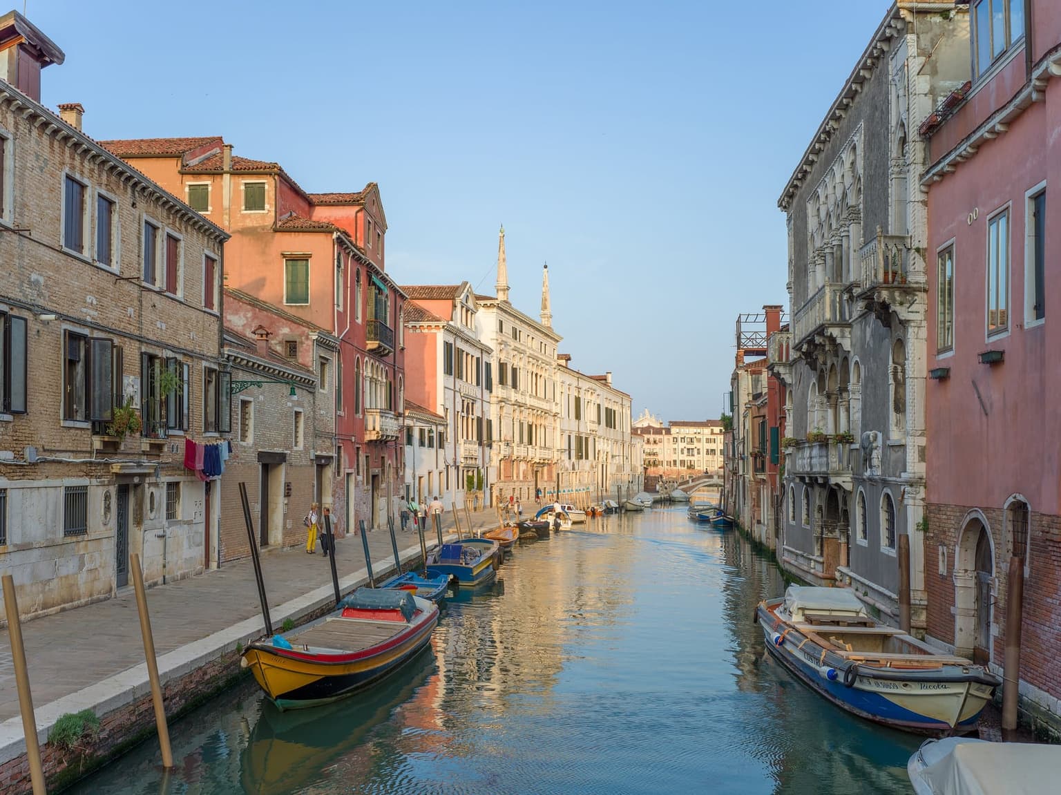 A quiet canal in Cannaregio lined by a long stone fondamenta, with laundry hanging from a brick house, moored wooden boats, a Gothic palazzo at right and a footbridge at the far end