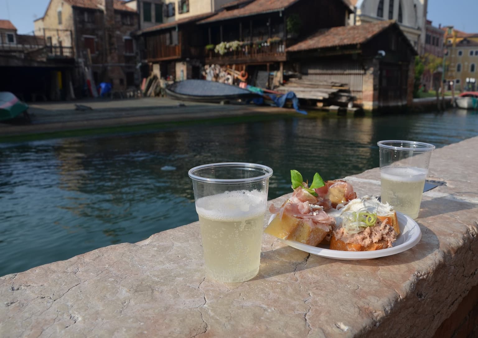 Two plastic cups of prosecco and a plate of cicchetti, bread topped with prosciutto and a creamy spread, set on a stone canal wall across from the wooden boatsheds of the San Trovaso gondola yard