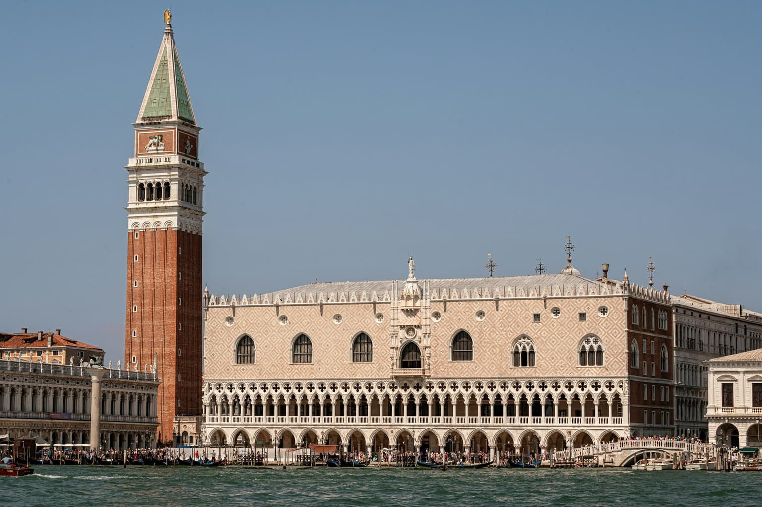 The Doge's Palace waterfront facade, its pink-and-white patterned stone above two tiers of Gothic arcades, seen from the water, with the brick Campanile of San Marco rising at left and crowds along the quay