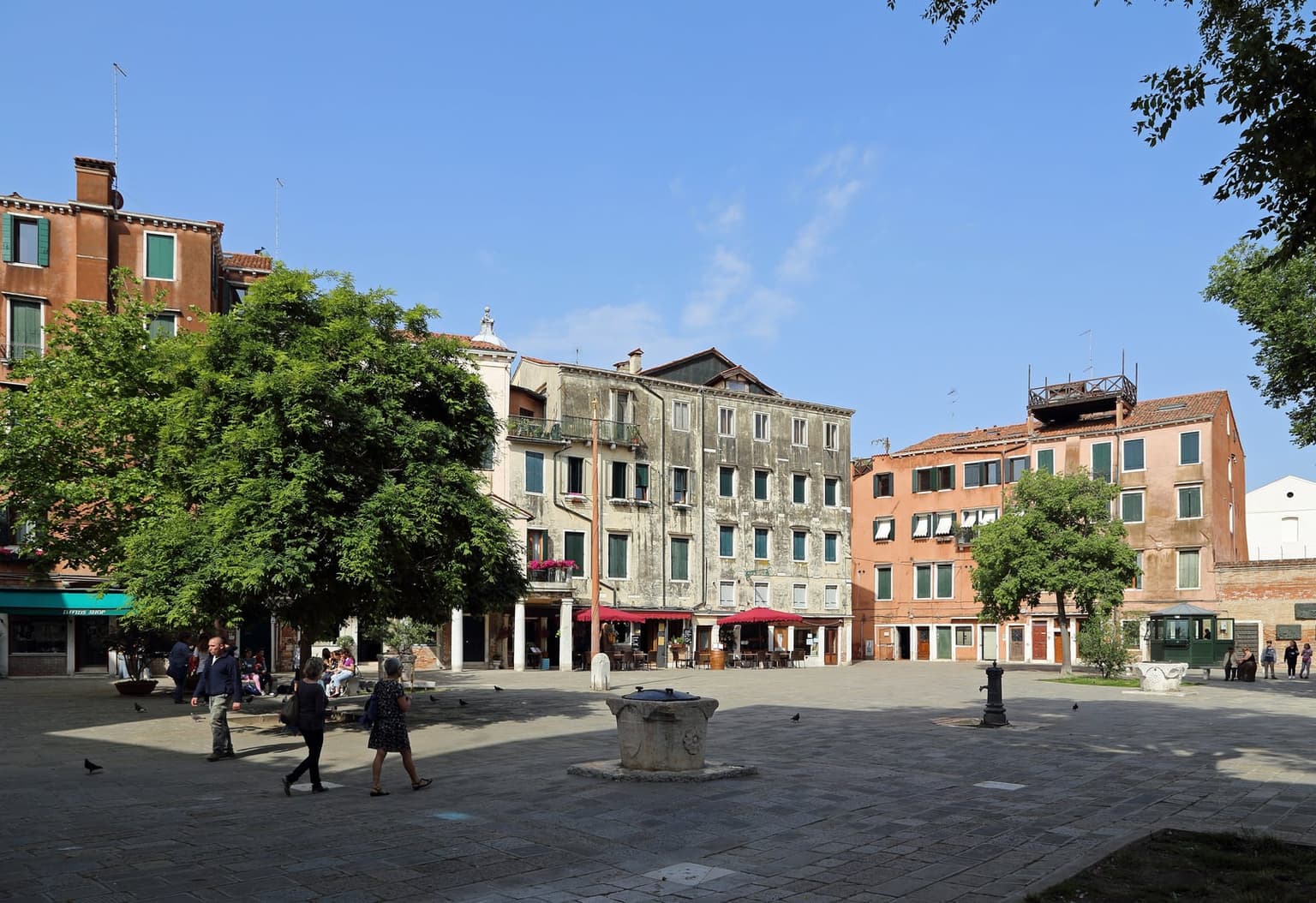 Campo del Ghetto Nuovo in Cannaregio on a sunny day, a stone wellhead in the middle of the square, tall plain houses with shops and café umbrellas at their base, trees and a few people walking