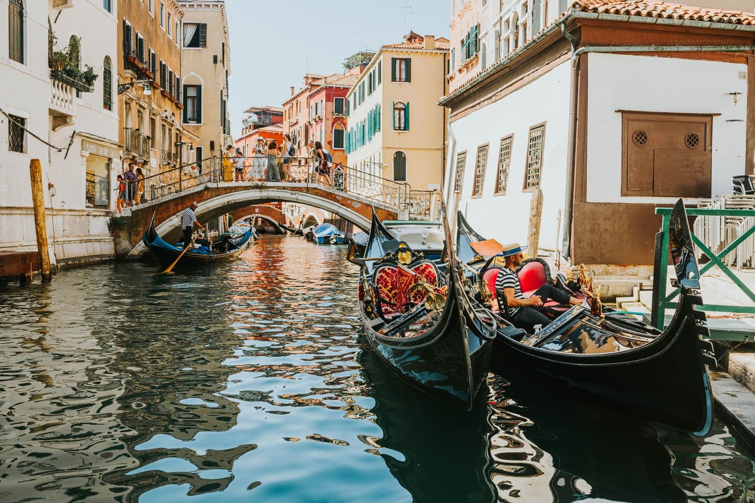 Two black gondolas with red-and-gold upholstered seats moored along a narrow Venice canal, a gondolier in a straw boater seated in one, while another gondolier rows beneath a stone footbridge crowded with sightseers