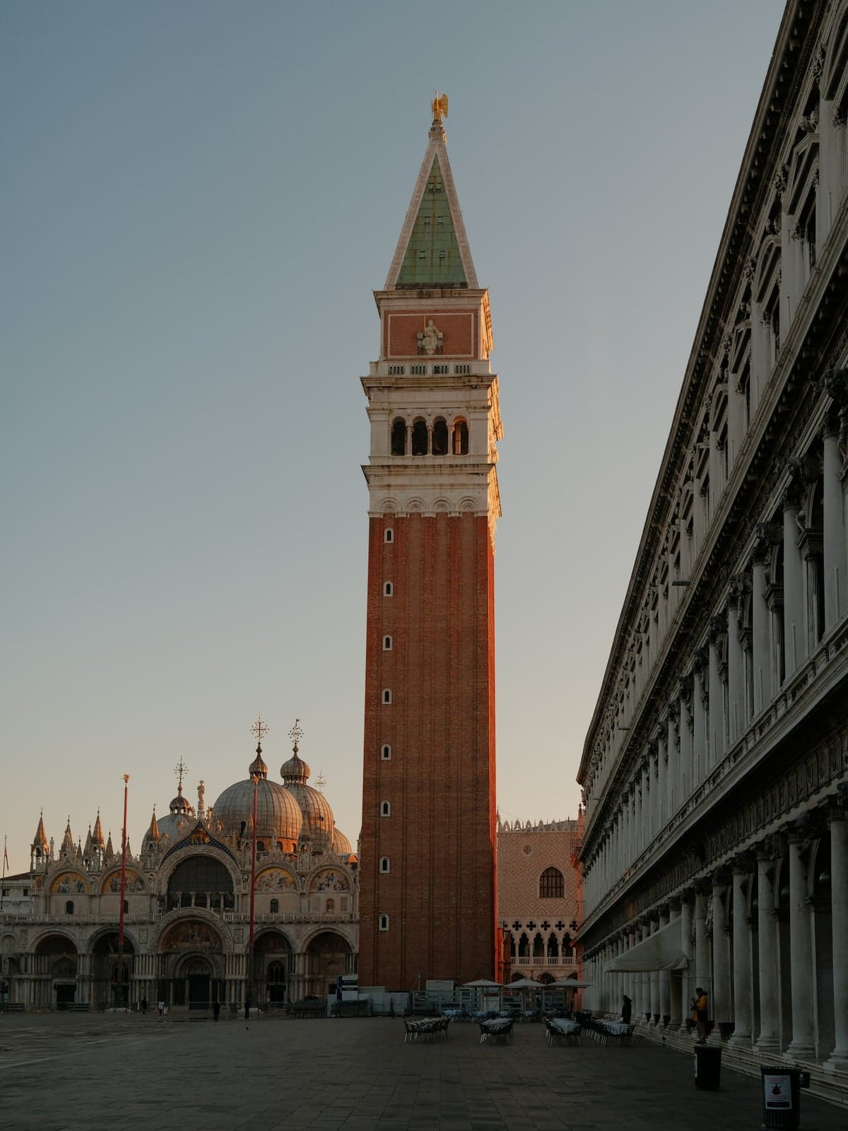Piazza San Marco almost empty in early morning light, the Campanile rising beside the domes and facade of St. Mark's Basilica, café tables set out and a few people under the arcades