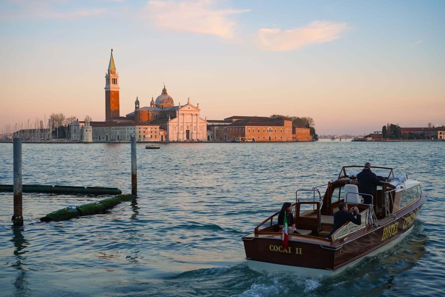 The church and bell tower of San Giorgio Maggiore across the water at sundown under a pink sky, with a varnished wooden motorboat crossing in the foreground