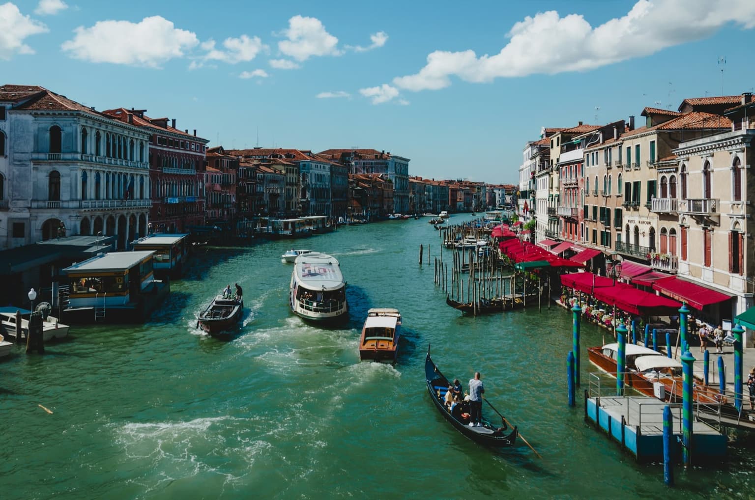 A vaporetto waterbus heading down the Grand Canal among a gondola, water taxis and a motorboat, seen from the Rialto Bridge, with floating vaporetto stops at left and red restaurant awnings along the right bank