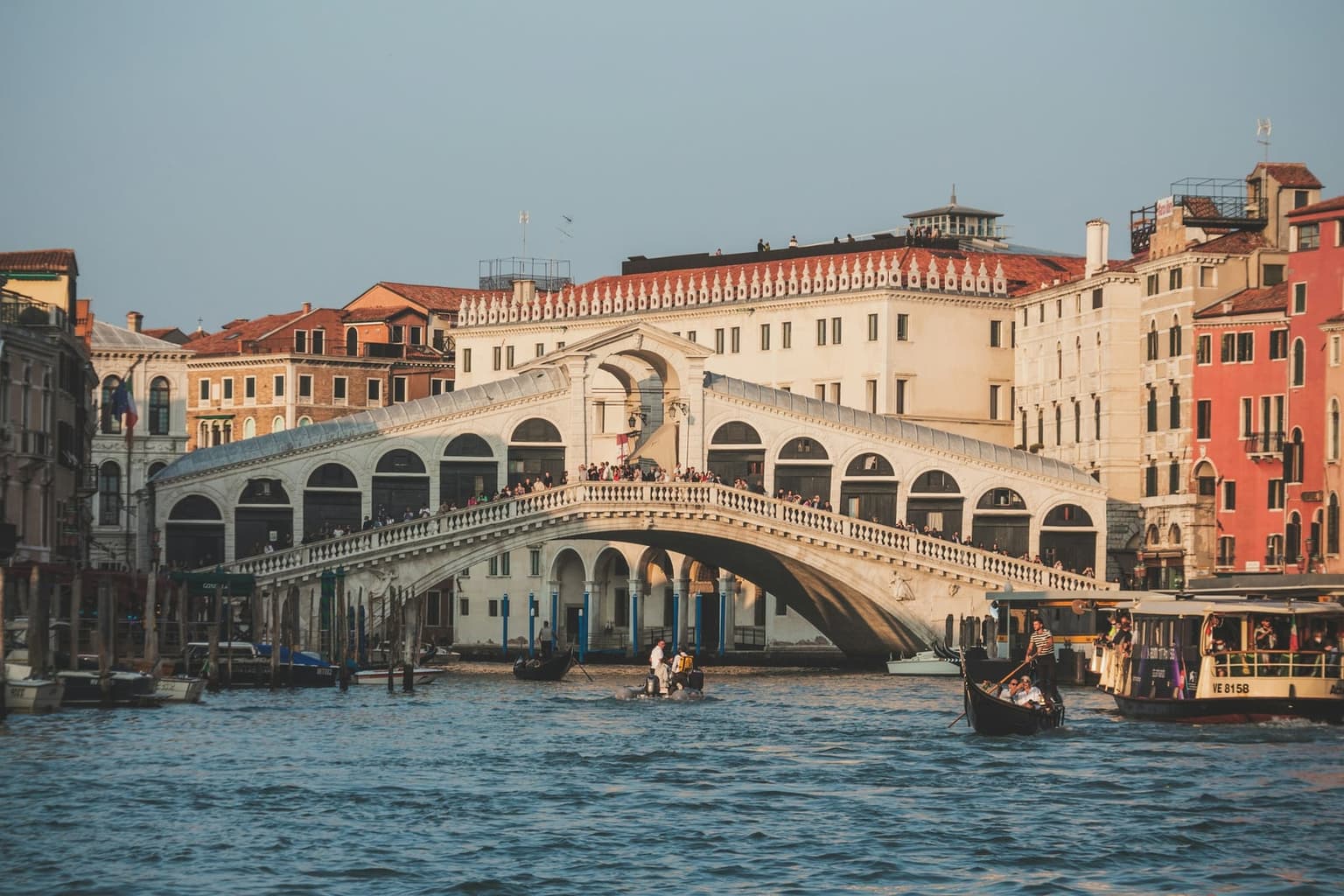 The Rialto Bridge over the Grand Canal seen from the water in warm low sun, people lining its balustrade, with a gondola, a small motorboat and a vaporetto passing below the palazzi