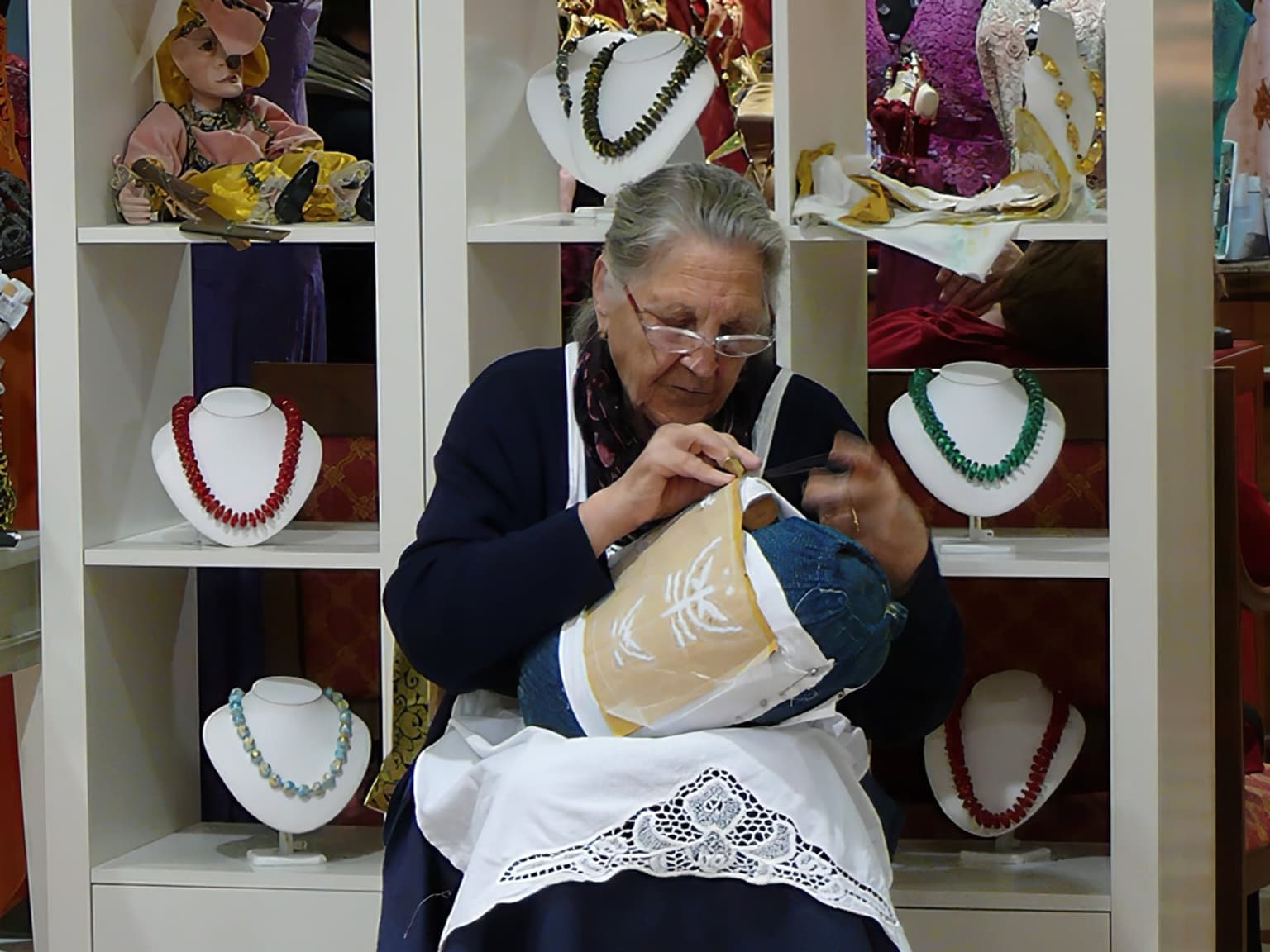 An elderly lacemaker in glasses and a white lace-trimmed apron working needle lace over a pattern on a round pillow in her lap, with shelves of necklaces on display behind her