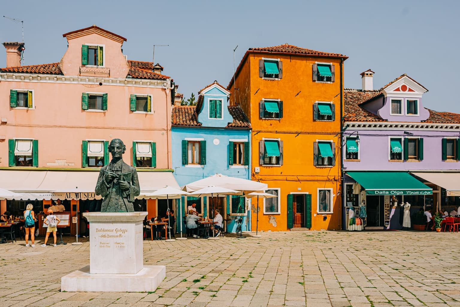 Piazza Galuppi on Burano, with the bronze bust of the composer Baldassare Galuppi on a white stone plinth in front of pink, blue, orange and lilac houses, café umbrellas and a lace shop under a green awning