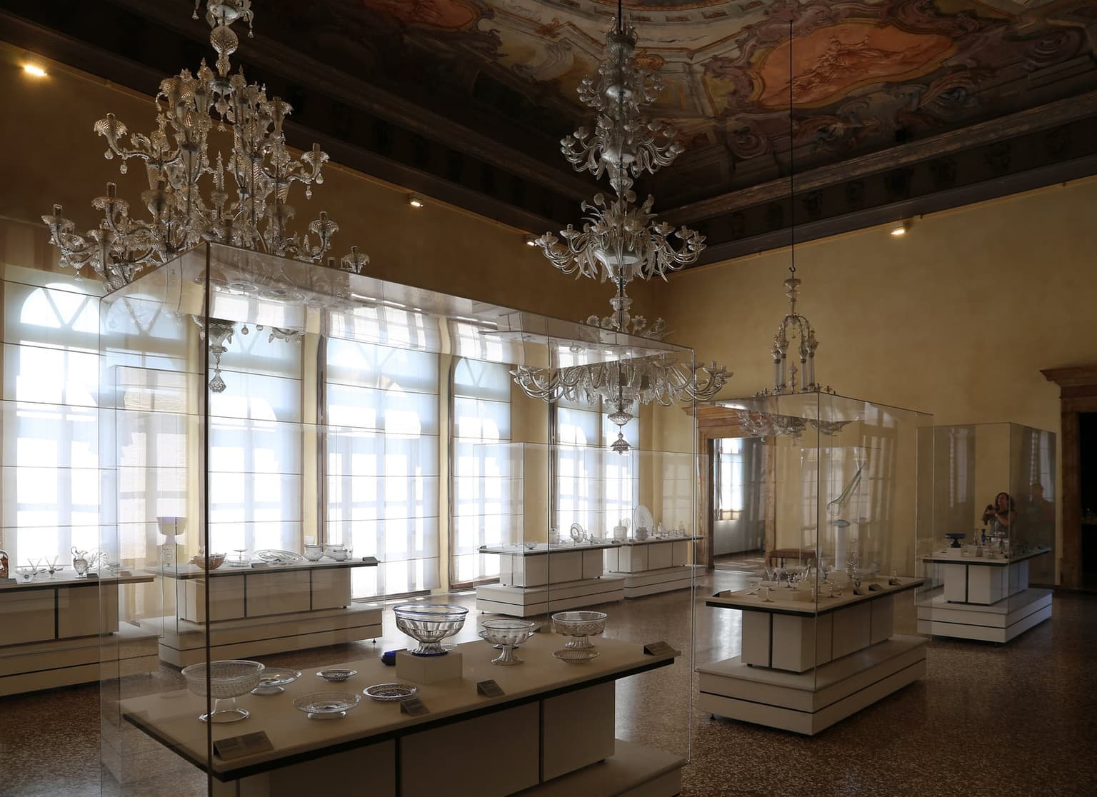 A frescoed hall in the Glass Museum on Murano, hung with large clear glass chandeliers, its display cases holding historic glass bowls, goblets and dishes