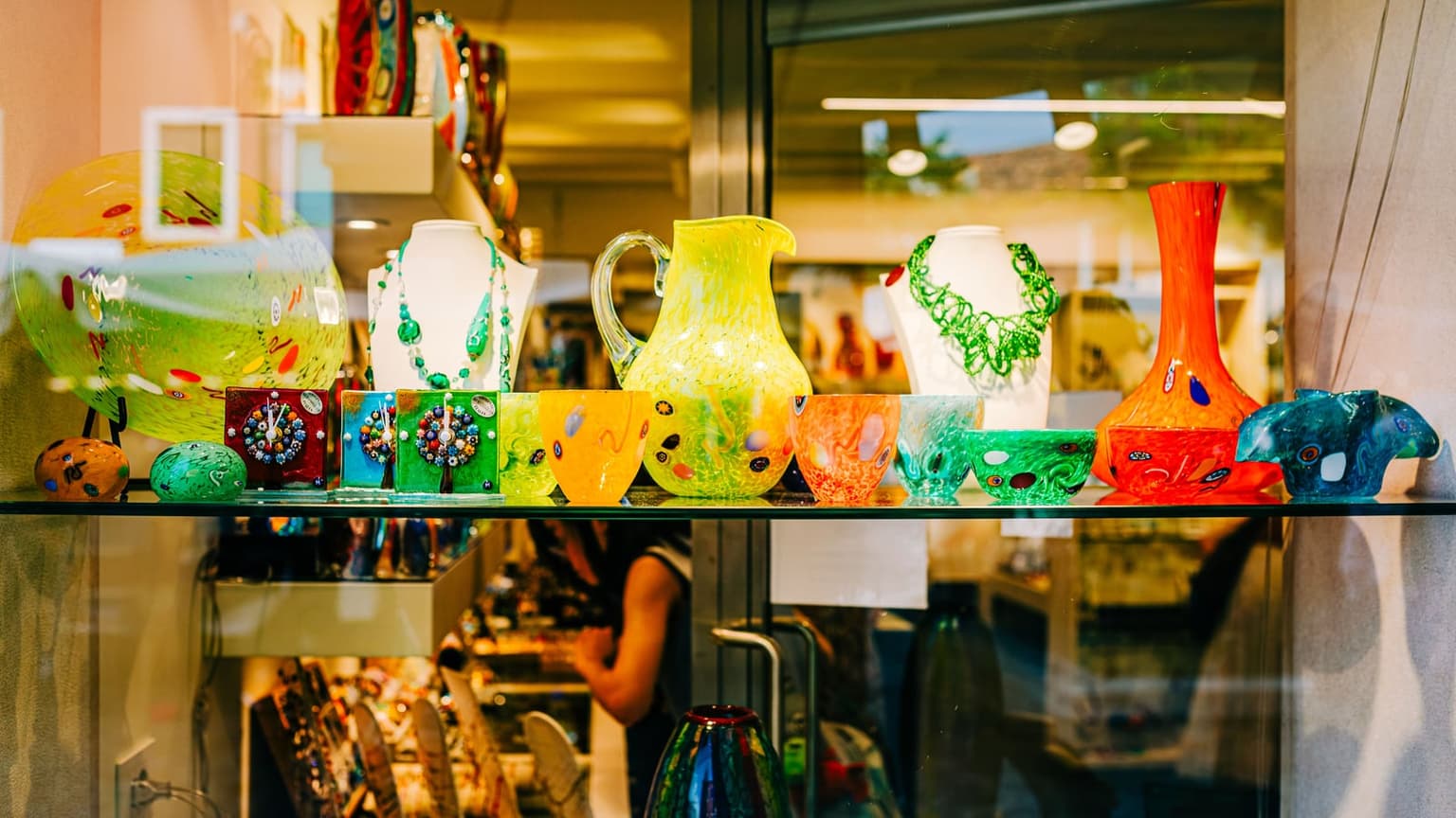 A shop window shelf of brightly colored glass, including a yellow speckled pitcher, millefiori-patterned cups and small clocks, a tall red vase and green glass-bead necklaces on display busts