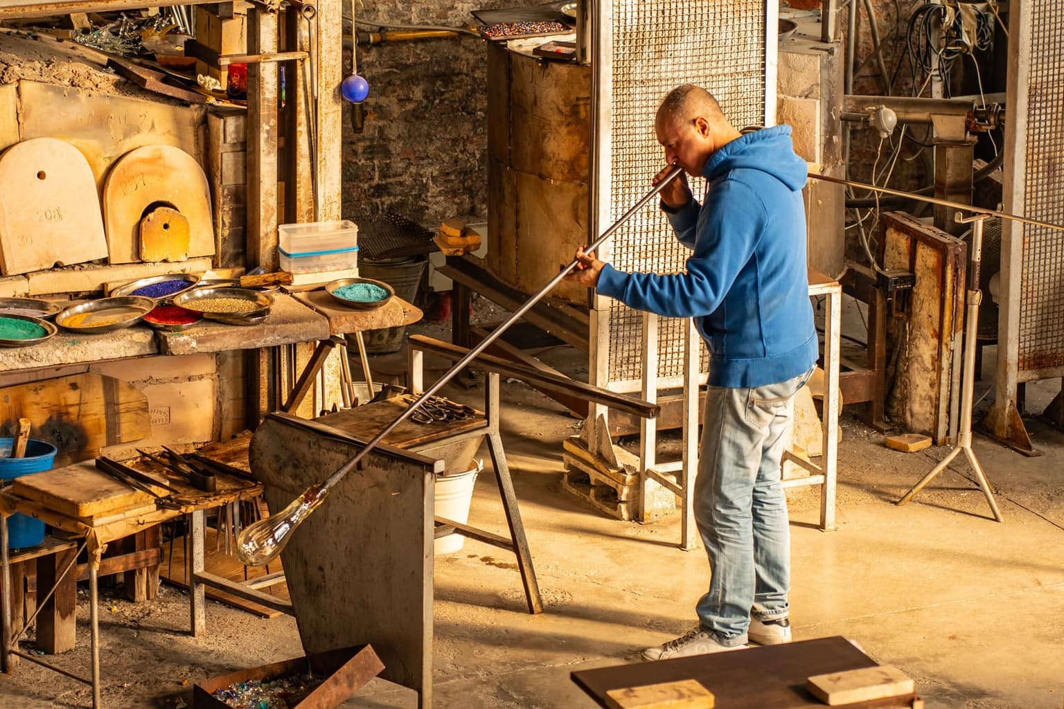 A glassblower blowing through a long iron blowpipe in a furnace workshop, a clear glass bubble forming at the far end, with bowls of colored glass granules and furnace doors behind him