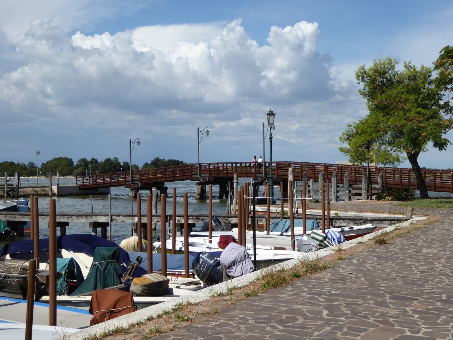 A long wooden footbridge with brown railings and lamp posts crossing a stretch of lagoon to a low, tree-lined shore, with small motorboats moored along a paved waterside path in the foreground