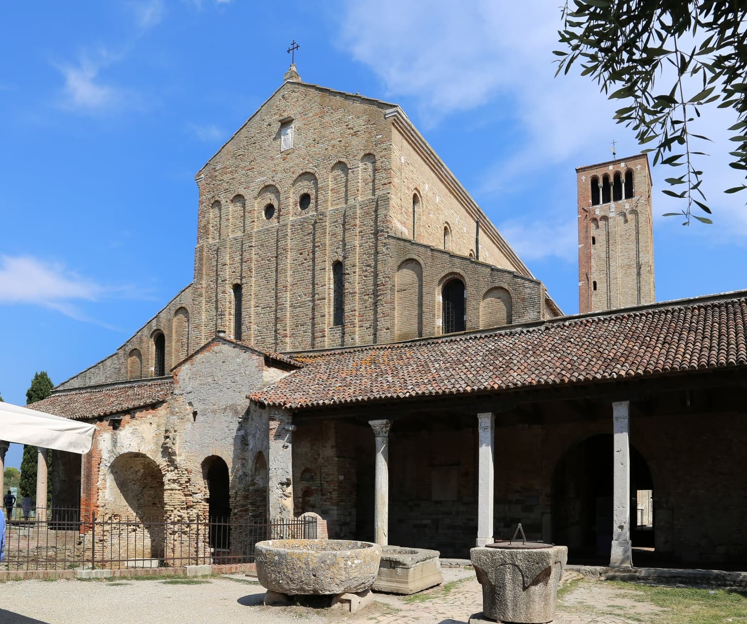 The tall brick front of the basilica on Torcello with its square brick bell tower rising at right, a tiled portico on stone columns and old stone wellheads in the gravel yard in front