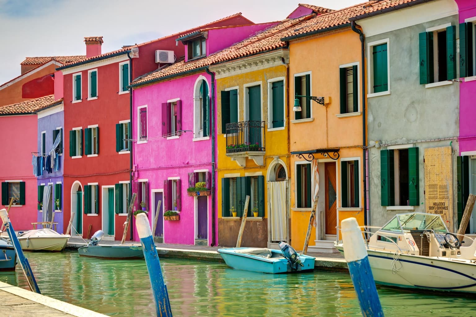 A row of brightly painted houses in red, pink, yellow and orange along a canal on Burano, with green shutters, laundry hanging from a window and small motorboats moored below blue and white mooring poles
