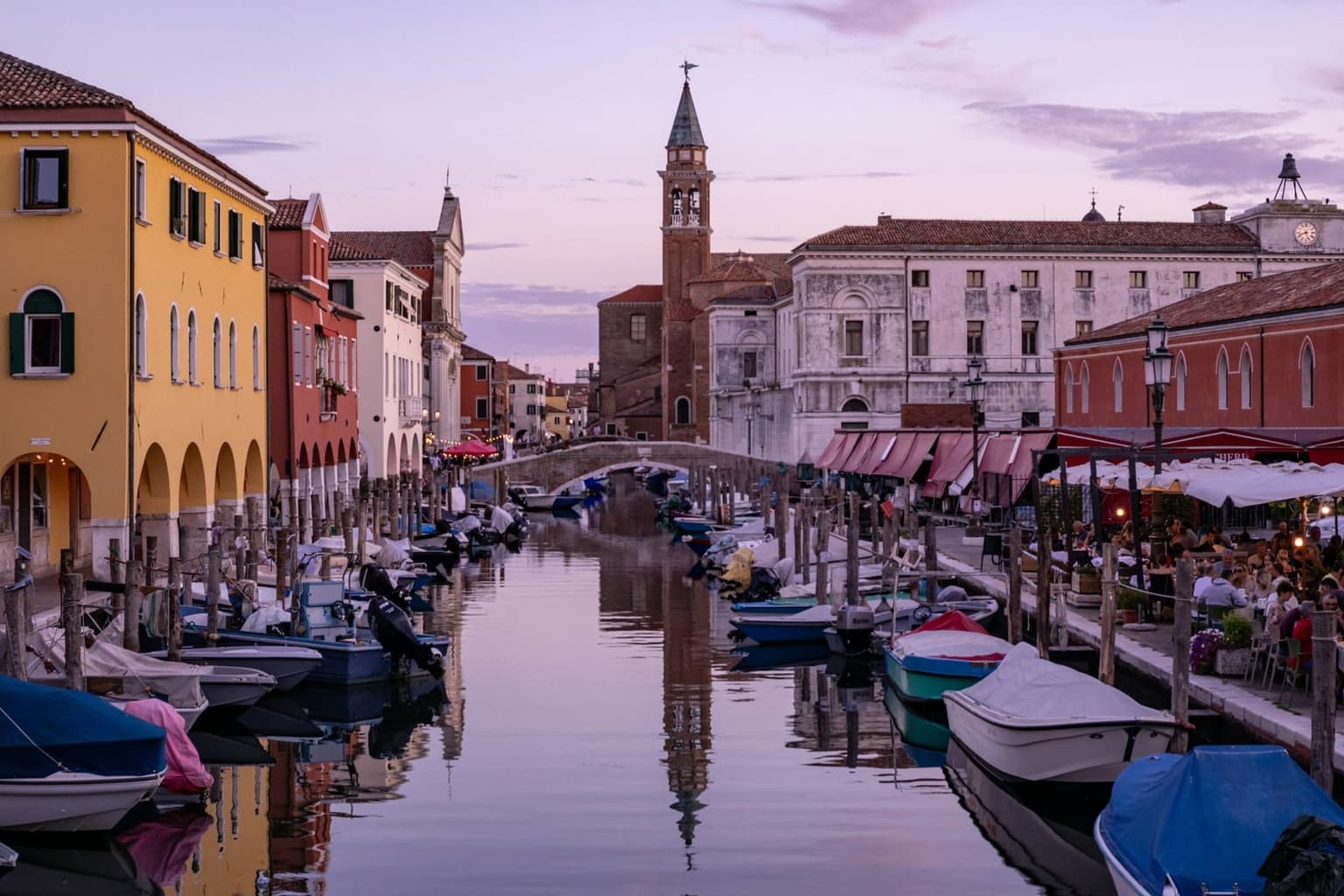 A canal in Chioggia at dusk lined with small moored motorboats, a yellow arcaded building on the left and restaurant tables under red awnings on the right, with a stone footbridge and a brick bell tower at the far end reflected in the still water