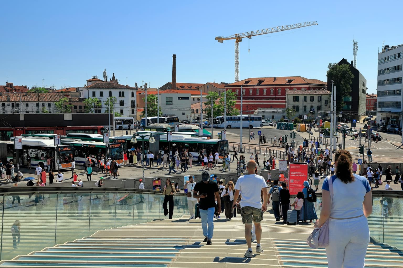 The Piazzale Roma bus terminal in Venice seen from the glass steps of the Ponte della Costituzione, with city buses lined up at numbered bays, travelers rolling suitcases down the steps, and a red sign for Venice's city access fee