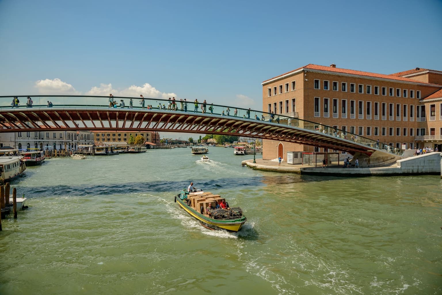 The Ponte della Costituzione, a shallow red steel and glass arch footbridge, spanning the Grand Canal with people crossing, a cargo boat stacked with boxes in the foreground and vaporetto stops along the left bank