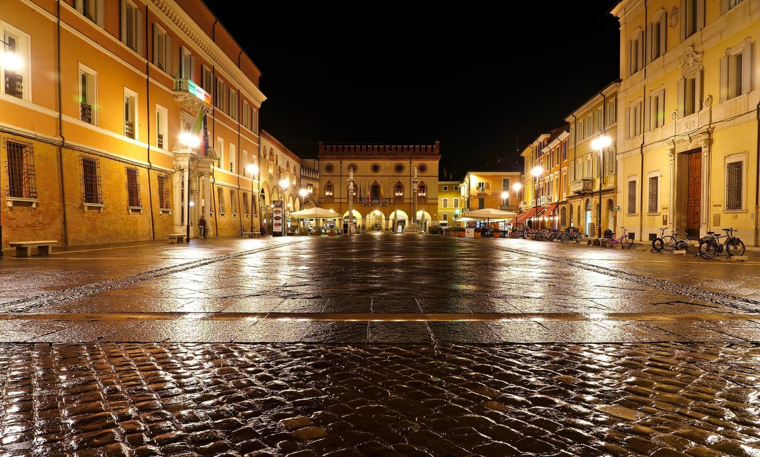 Ravenna's Piazza del Popolo at night after rain, wet paving reflecting the streetlights, with the crenellated Palazzo Comunale and its two stone columns at the far end and cafe umbrellas and parked bicycles along the sides